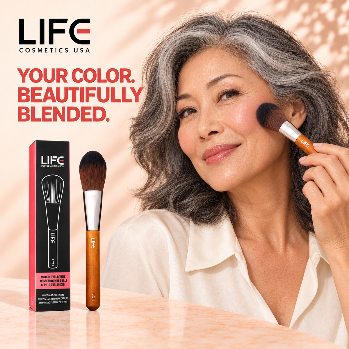 Woman with wavy gray hair applying makeup with the LIFE Cosmetics USA LC11 Medium Oval Brush, beside the brush and retail box.