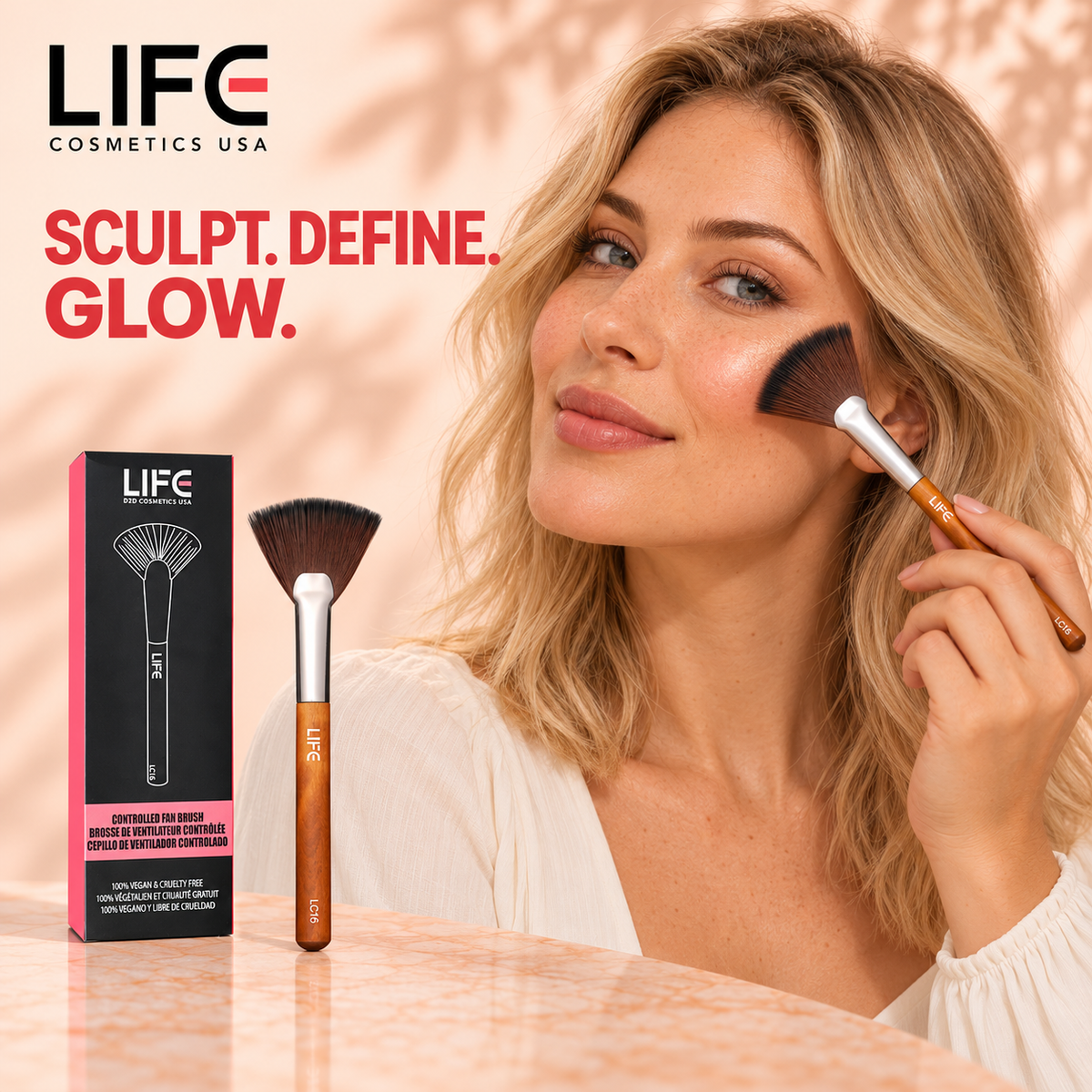 Blonde woman applying makeup to her cheek with the LIFE Cosmetics USA LC16 Controlled Fan Brush, beside the brush and retail box.