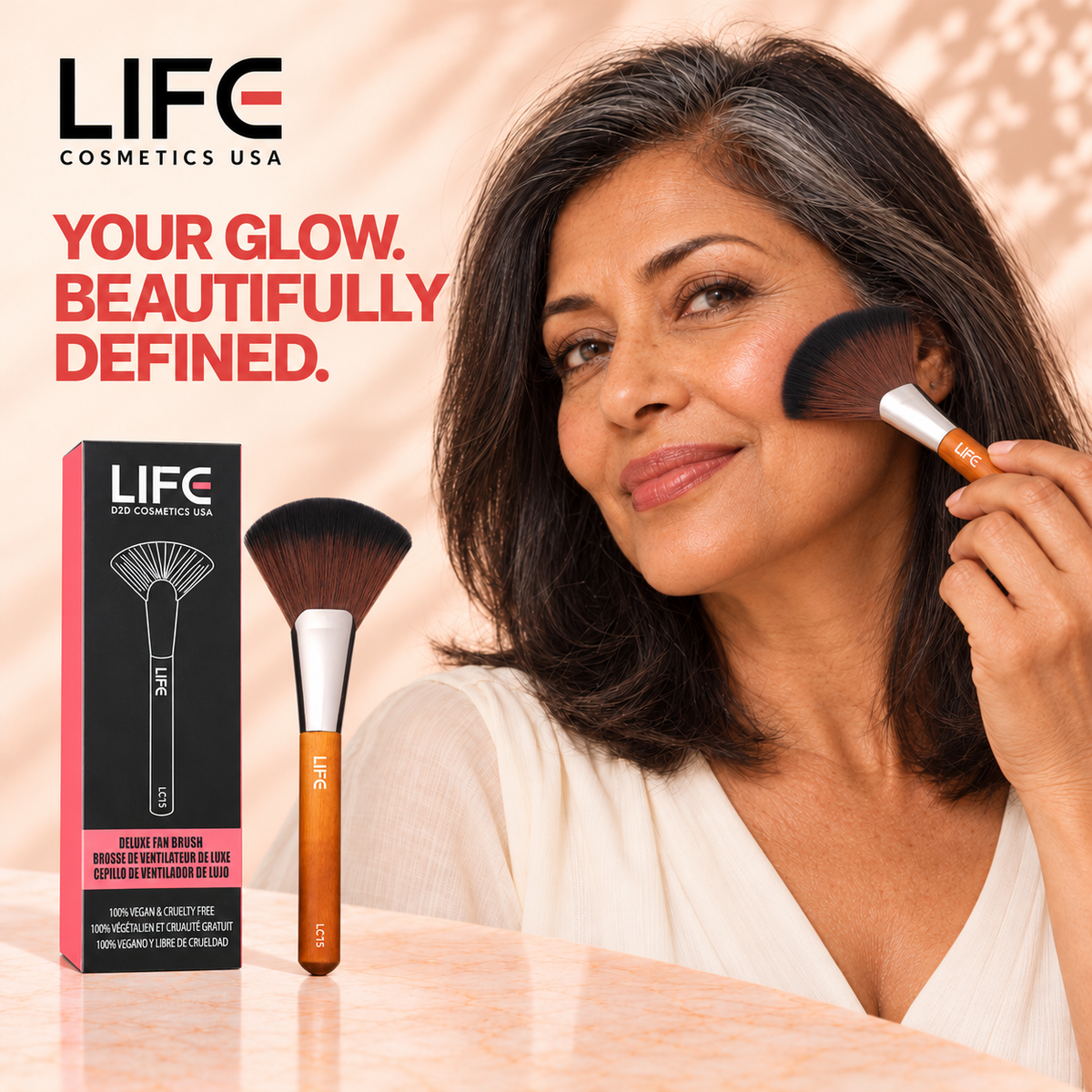 Woman with shoulder-length dark and gray hair using the LIFE Cosmetics USA LC15 Deluxe Fan Brush on her cheek, beside its retail box.
