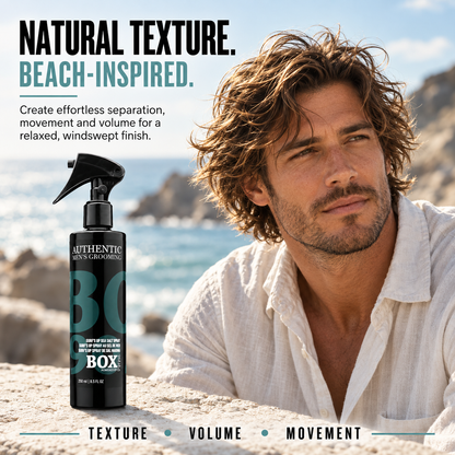 Man with tousled wavy hair wearing a white shirt beside a BOX90210 Surf’s Up Sea Salt Spray bottle on a stone ledge overlooking the ocean.
