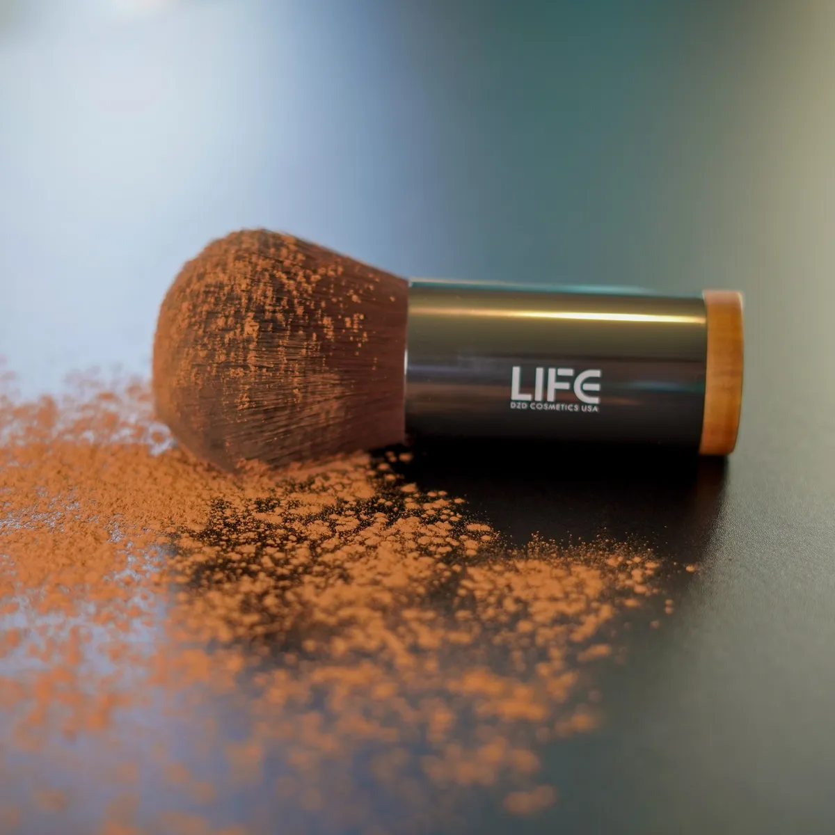Makeup brush with LIFE brand on a reflective surface with scattered powder.
