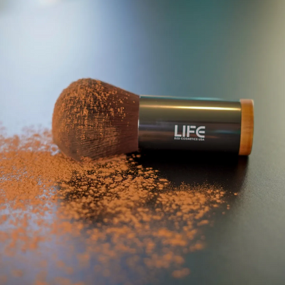 Makeup brush with LIFE brand on a reflective surface with scattered powder.